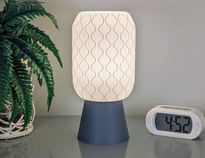 A Cirrus Table Lamp from Dryft Home, featuring a 3D printed airy elegance with a wavy pattern and dark base, casts its glow beside a digital clock displaying 4:52. Fern leaves introduce greenery to the minimalist scene, highlighting sustainable materials in harmonious balance.