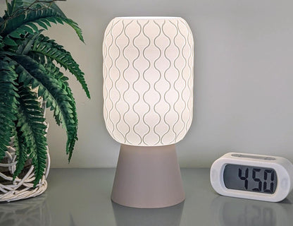 A Cirrus Table Lamp, characterized by its 3D printed airy elegance, gently illuminates a table. Alongside, a digital clock showing 4:50 and a potted fern in a woven basket enhance this modern design from Dryft Home, beautifully crafted from sustainable materials.