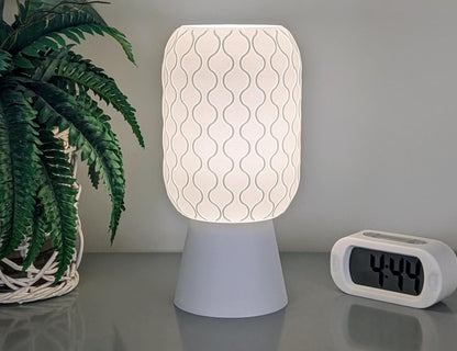 The Cirrus Table Lamp – 3D Printed Airy Elegance by Dryft Home is crafted from sustainable materials and showcases a white wavy patterned shade. It emits a gentle glow onto a surface near a digital clock displaying 4:44 and a potted fern, creating a serene, ambient atmosphere.