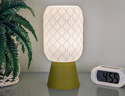 A patterned, glowing Cirrus Table Lamp – 3D Printed Airy Elegance by Dryft Home with a green base sits on a surface next to a potted plant. An alarm clock displaying the time 4:55 is on the right.