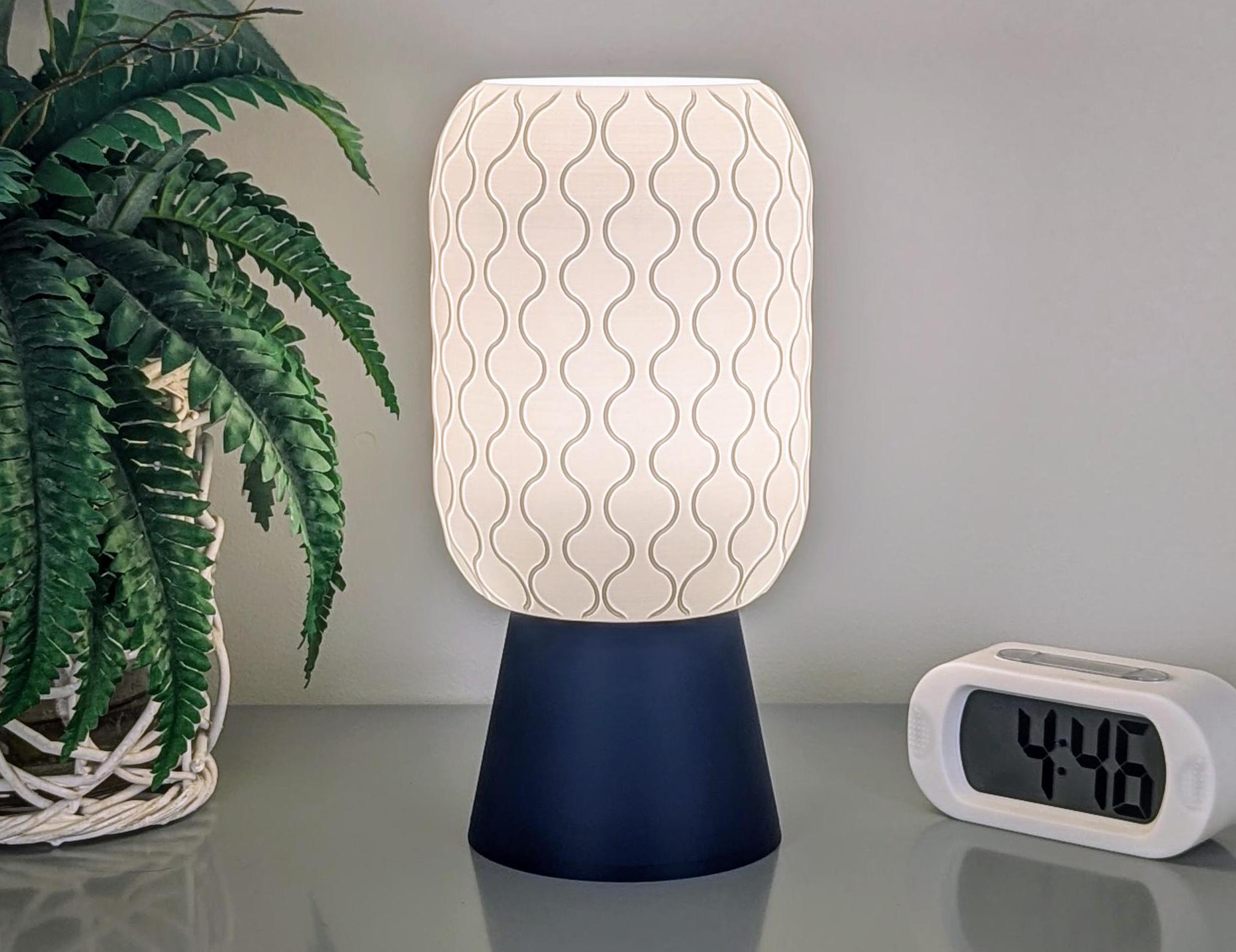 The Cirrus Table Lamp by Dryft Home, featuring a wavy patterned shade crafted from sustainable materials and a dark base, is illuminated on a desk. Alongside it, a digital clock showing 4:48 and a potted fern contribute to an elegant and eco-friendly ambiance.