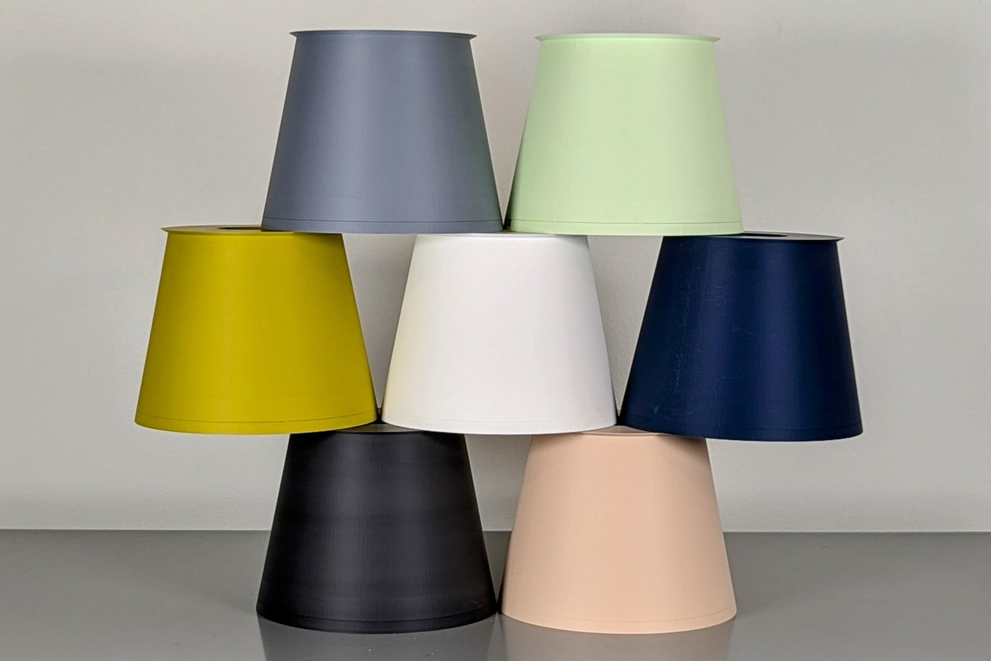 A collection of vibrant Cirrus Table Lamps by Dryft Home, showcasing a pyramid arrangement of conical shades made from sustainable materials. The shades are available in gray, lime, pale green, white, cream, black, and navy blue, all displayed against a simple gray backdrop.