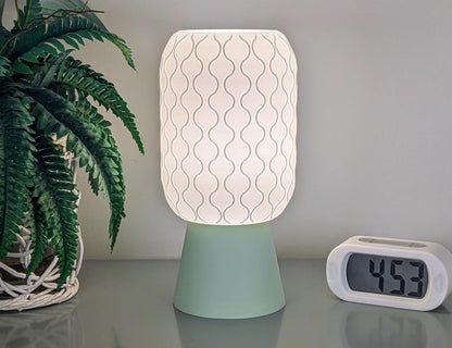 A decorative Cirrus Table Lamp – 3D Printed Airy Elegance by Dryft Home, featuring a wavy pattern on its shade, is elegantly perched on a light green base. It sits beside a potted plant and a digital clock displaying 4:59, showcasing its design crafted from sustainable materials.