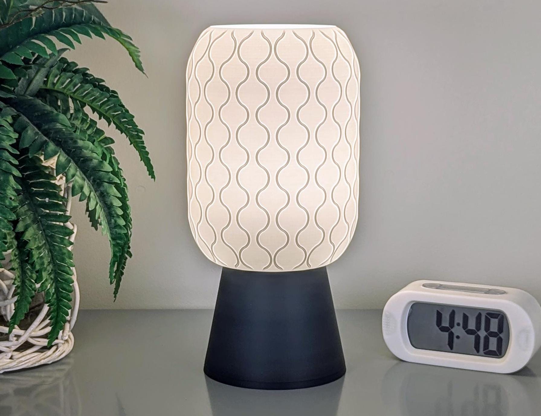 A modern Cirrus Table Lamp – 3D Printed Airy Elegance by Dryft Home, featuring a wavy pattern on its white shade, rests on a black base. Beside it, a digital clock displays the time as 4:48. A nearby potted plant with green leaves contributes a natural element to the sleek design, which is crafted from sustainable materials.