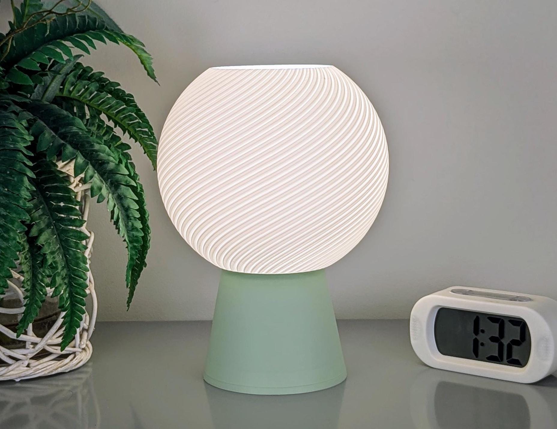 The Vortice Table Lamp by Dryft Home, featuring a textured, glowing spherical shade created with sustainable 3D printing techniques, is elegantly perched on a light green base. It rests on a gray surface beside a white digital clock displaying 1:32 and a potted plant with lush green leaves in a woven basket.