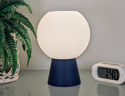 The Vortice Table Lamp by Dryft Home, a contemporary lighting piece with a spherical, textured white shade made from biodegradable PLA plastic, sits on a reflective surface. To the right of this sustainable creation is a digital clock displaying 1:40. On the left, a potted plant with vibrant green leaves adds to the aesthetic appeal.