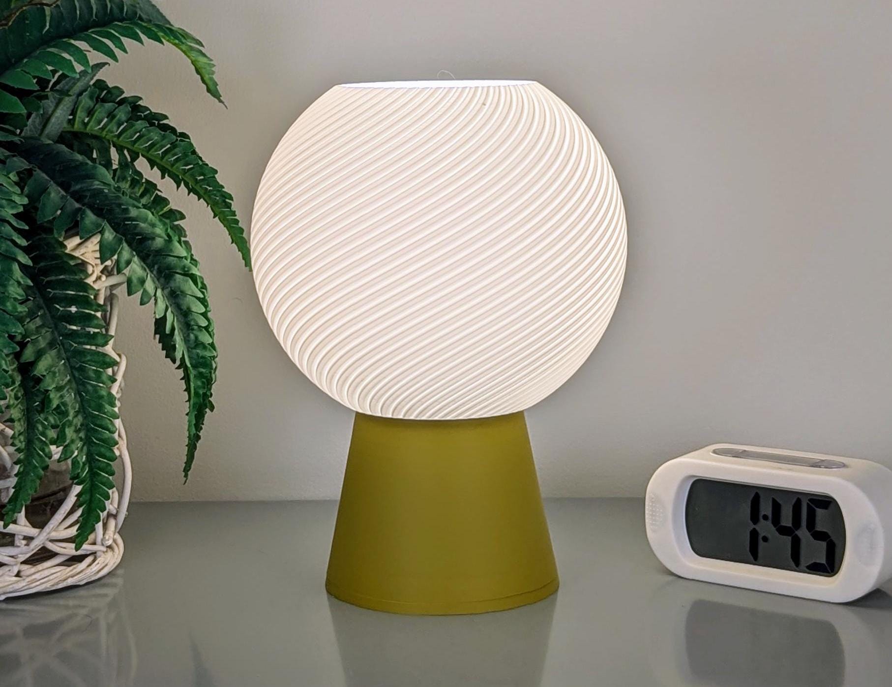 A Vortice Table Lamp by Dryft Home, featuring a white, spherical design with spiral patterns and crafted from biodegradable PLA plastic, rests on a green base. Nearby is a digital clock displaying 1:45. To the left of this modern lighting piece stands a potted fern, adding a touch of natural elegance to the setting.