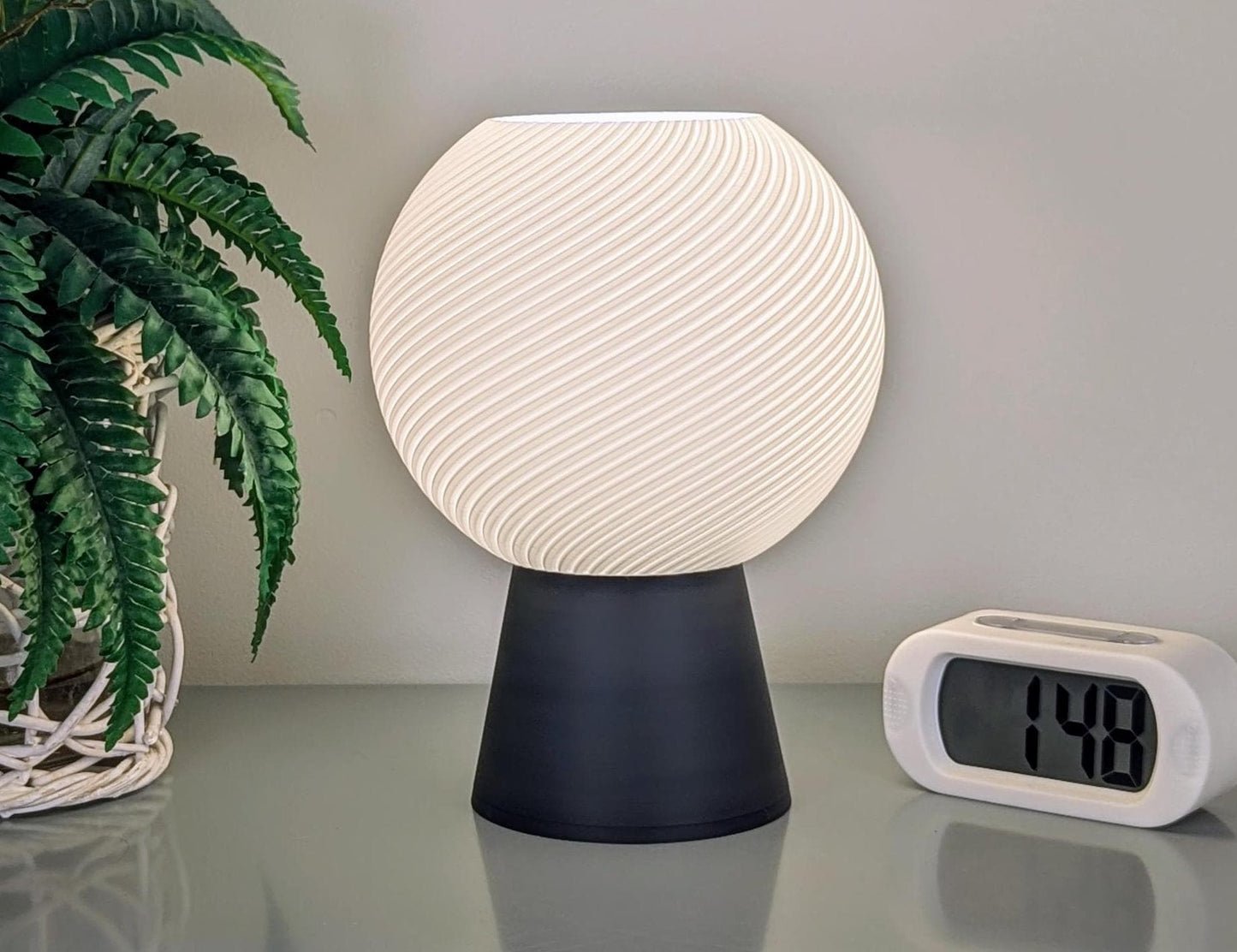 The Vortice Table Lamp by Dryft Home, featuring a spherical design with a textured white shade made from biodegradable PLA plastic, rests on a dark base beside a digital clock displaying 1:48 on a sleek surface. To the left, there's a potted plant with green leaves, underscoring the dedication to sustainable production.