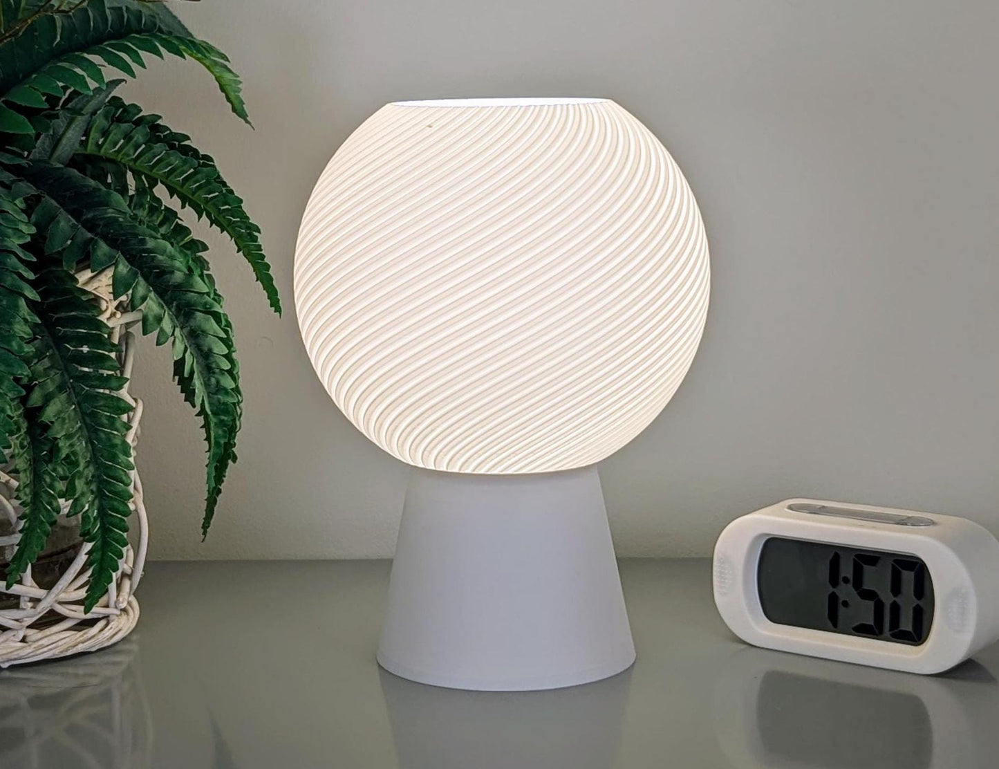 The Vortice Table Lamp by Dryft Home, crafted from biodegradable PLA plastic, casts a gentle light over the table. Nearby, a digital clock shows 1:50, while a potted fern enhances the atmosphere with its lush greenery—all embodying principles of sustainable production.