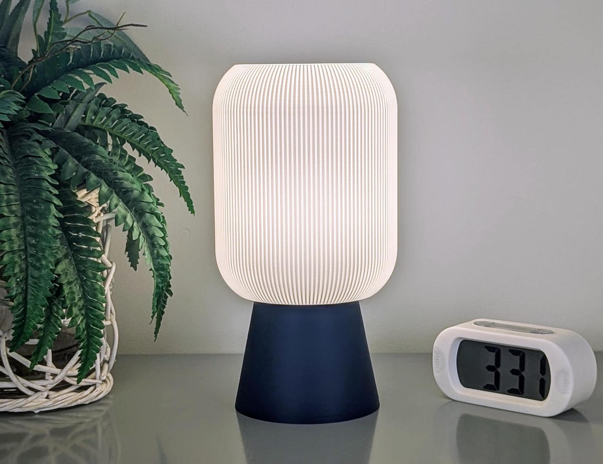 A Cumulis Table Lamp by Dryft Home, featuring a 3D printed pleated white shade and blue base, embraces modern design as it sits beside a digital clock showing 3:31. To its left, a potted fern with textured leaves adds a touch of nature against the plain, light-colored wall.