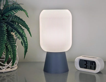 The Cumulis Table Lamp by Dryft Home, featuring a 3D printed pleated cylindrical shade made from sustainable materials, rests on a sleek dark base. It illuminates a reflective surface beside a digital clock displaying 3:14. A potted plant with vibrant green leaves adds an eco-conscious touch to the scene.