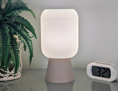 The Cumulis Table Lamp by Dryft Home, featuring a 3D-printed pleated cylinder design crafted from sustainable materials, emits a gentle glow on the gray surface. Next to it, a digital clock showing 3:27 and a potted fern in a woven basket together create an eco-friendly and cozy minimalist setting.