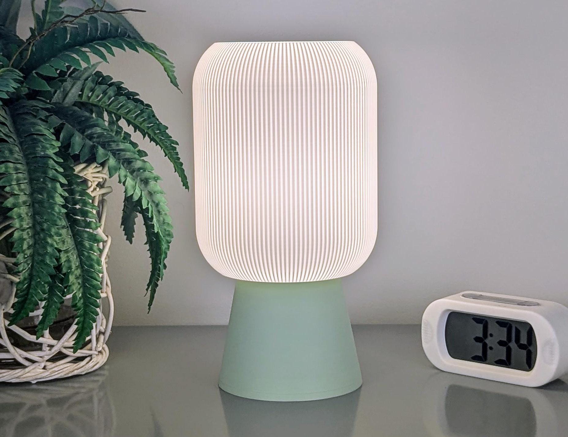 A Cumulis Table Lamp by Dryft Home, featuring a white, pleated shade and a green base crafted from sustainable materials, sits on a reflective surface. Beside it is a digital clock showing 3:34. On the left side is an eco-conscious touch with a leafy plant in a white woven basket.