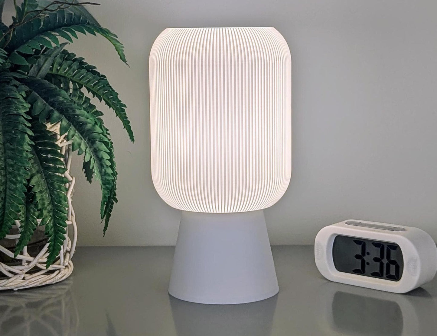 The Cumulis Table Lamp by Dryft Home, a 3D printed pleated cylinder design, casts a warm glow on a reflective surface. A white digital clock shows 3:36 to the right, with a potted plant featuring green leaves to the left.