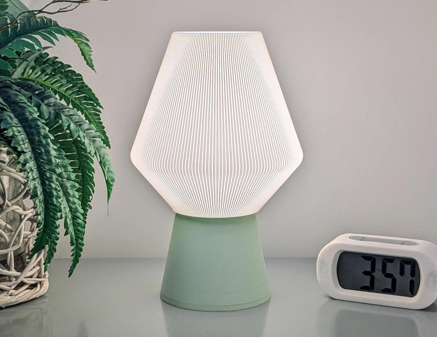 The Inversis Table Lamp from Dryft Home, crafted with sustainable materials, showcases a white geometric shade and green base. Positioned next to a potted fern and a digital clock displaying 3:59, it offers 3D printed sculptural lighting for a modern touch.