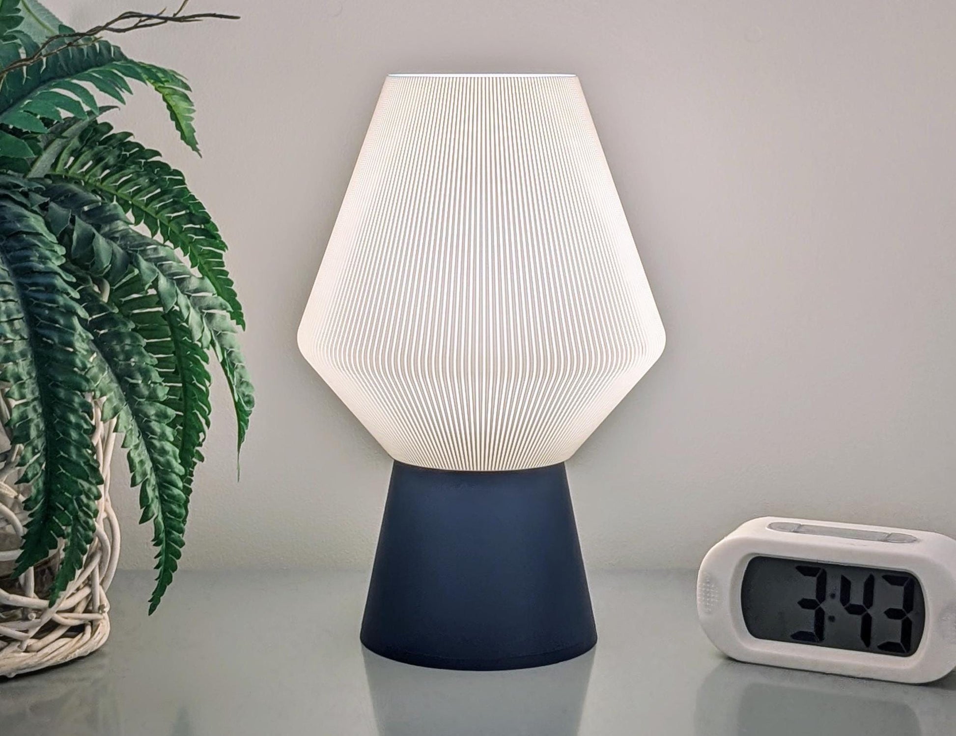 The Inversis Table Lamp by Dryft Home, with its sculptural and modern touch, features a textured white shade and dark base that rest elegantly on a reflective surface. To the left of this sustainably crafted lamp is a potted plant, while a digital alarm clock showing 3:49 sits to the right. It beautifully complements the simple, light-colored wall in the background.