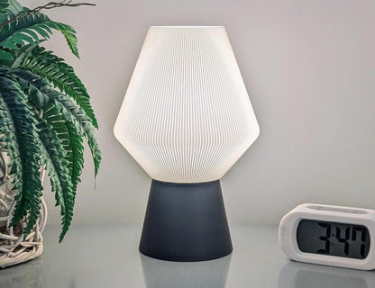 The Inversis Table Lamp from Dryft Home, made with sustainable materials, showcases a contemporary design featuring a white geometric shade paired with a dark base. Nearby, a digital clock shows the time as 3:47, while the green leaves of a potted plant are visible on the left side.