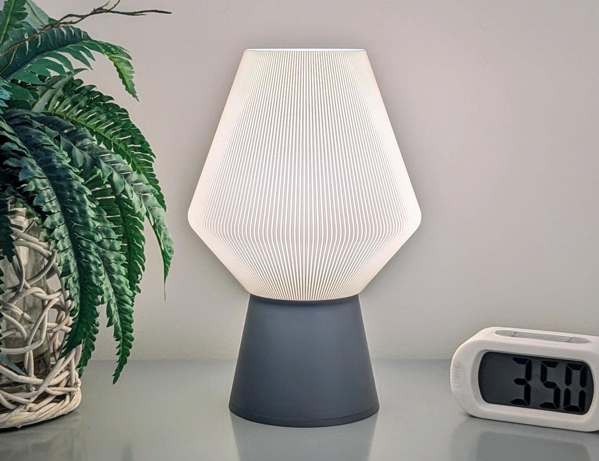 Positioned beside a digital clock reading 3:50 and a potted plant in a wicker basket, the Inversis Table Lamp by Dryft Home offers a sculptural, modern touch with its geometric design. This lamp, crafted from sustainable materials, emits a gentle glow on the desk.