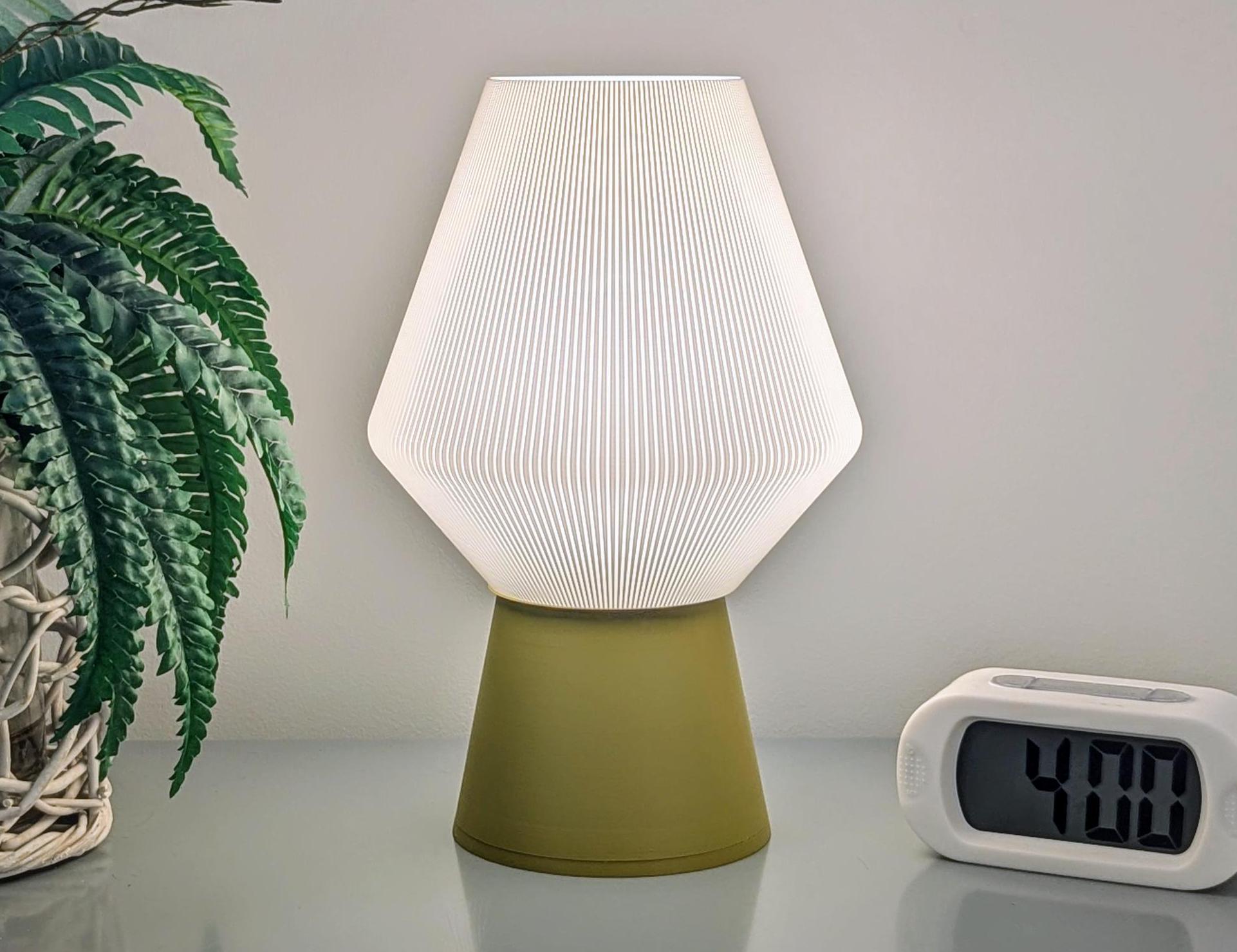 An Inversis Table Lamp by Dryft Home, featuring a modern design with a white pleated shade and a sustainably crafted green base, is placed on a surface beside a compact digital clock showing 4:00. On the left side, there is a potted plant with lush green foliage.