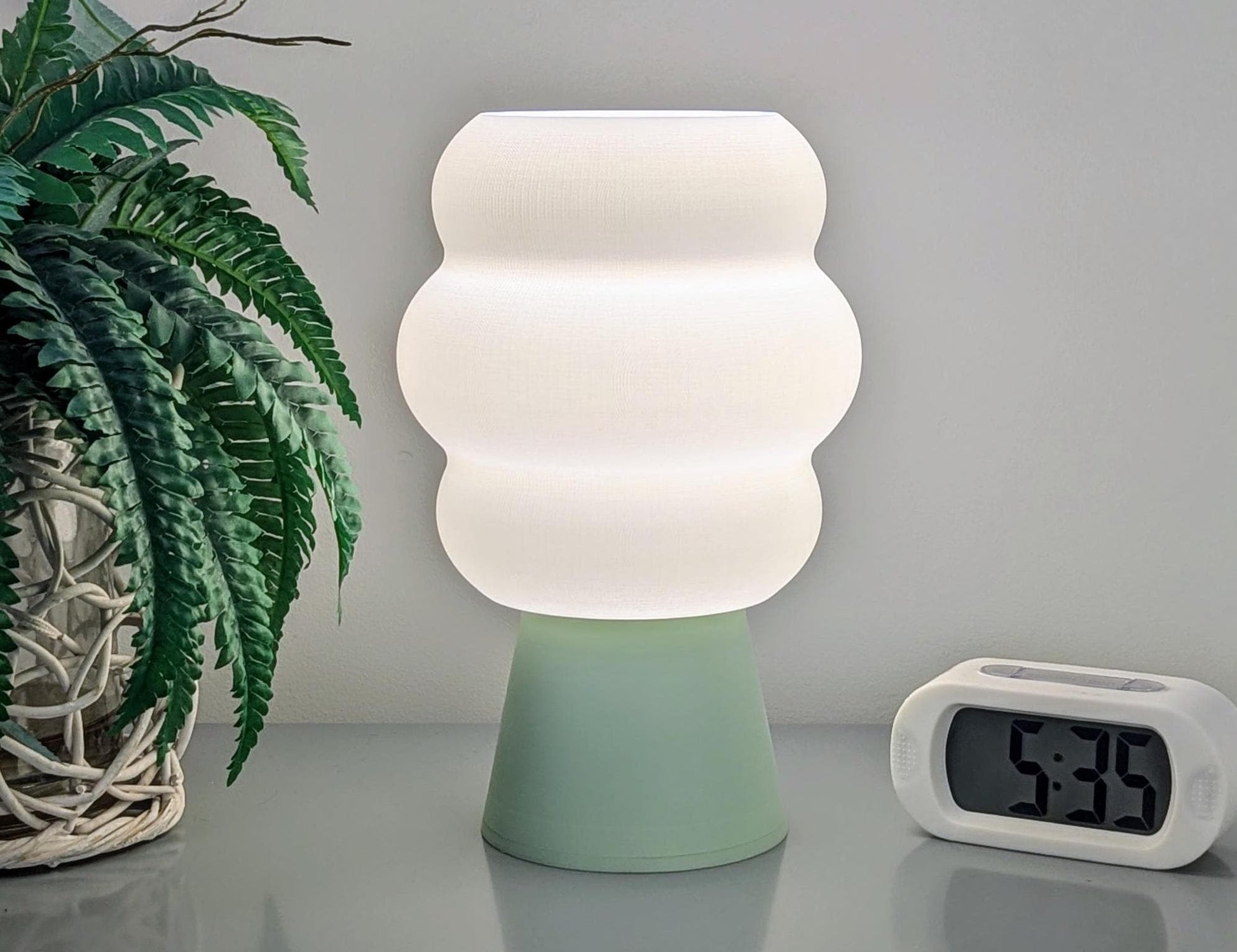 The Dryft Home Mammatus Table Lamp – 3D Printed Cloud-Inspired Lighting, featuring a modern design with a green base, illuminates a table next to a digital clock displaying 5:36. Crafted from biodegradable PLA plastic, it enhances the minimalist decor alongside a fern-like potted plant on the left, all arranged on a smooth, light-gray surface.