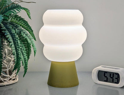 A Mammatus Table Lamp from Dryft Home, designed with biodegradable PLA plastic, boasts a contemporary look with its white wavy shade and yellow base. It sits beside a digital clock showing 5:49. To the left, a potted plant with lush green leaves complements this eco-friendly arrangement.