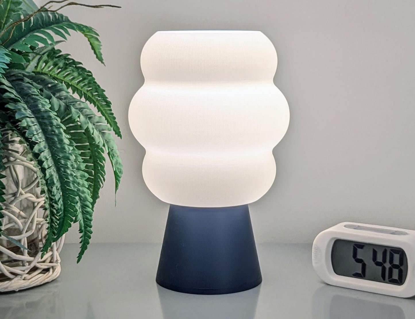 The Mammatus Table Lamp – 3D Printed Cloud-Inspired Lighting from Dryft Home, featuring a wavy, white shade crafted from biodegradable PLA plastic and a sleek black base, is elegantly placed on the table. Beside it sits a digital clock showing 5:48, while on the left side, a green fern in a woven basket creates a contrasting pop of color against the neutral background.