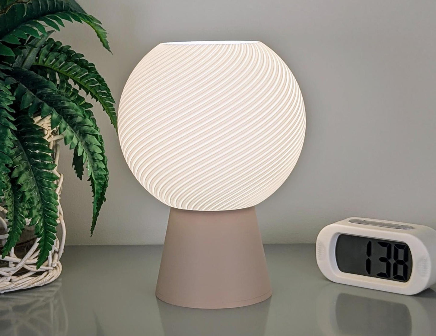 The Vortice Table Lamp by Dryft Home, featuring a spiraled globe shade and made from eco-friendly biodegradable PLA plastic, emits a gentle light from its beige base. It rests beside a digital alarm clock displaying 1:38 and a woven pot containing a vibrant green plant.