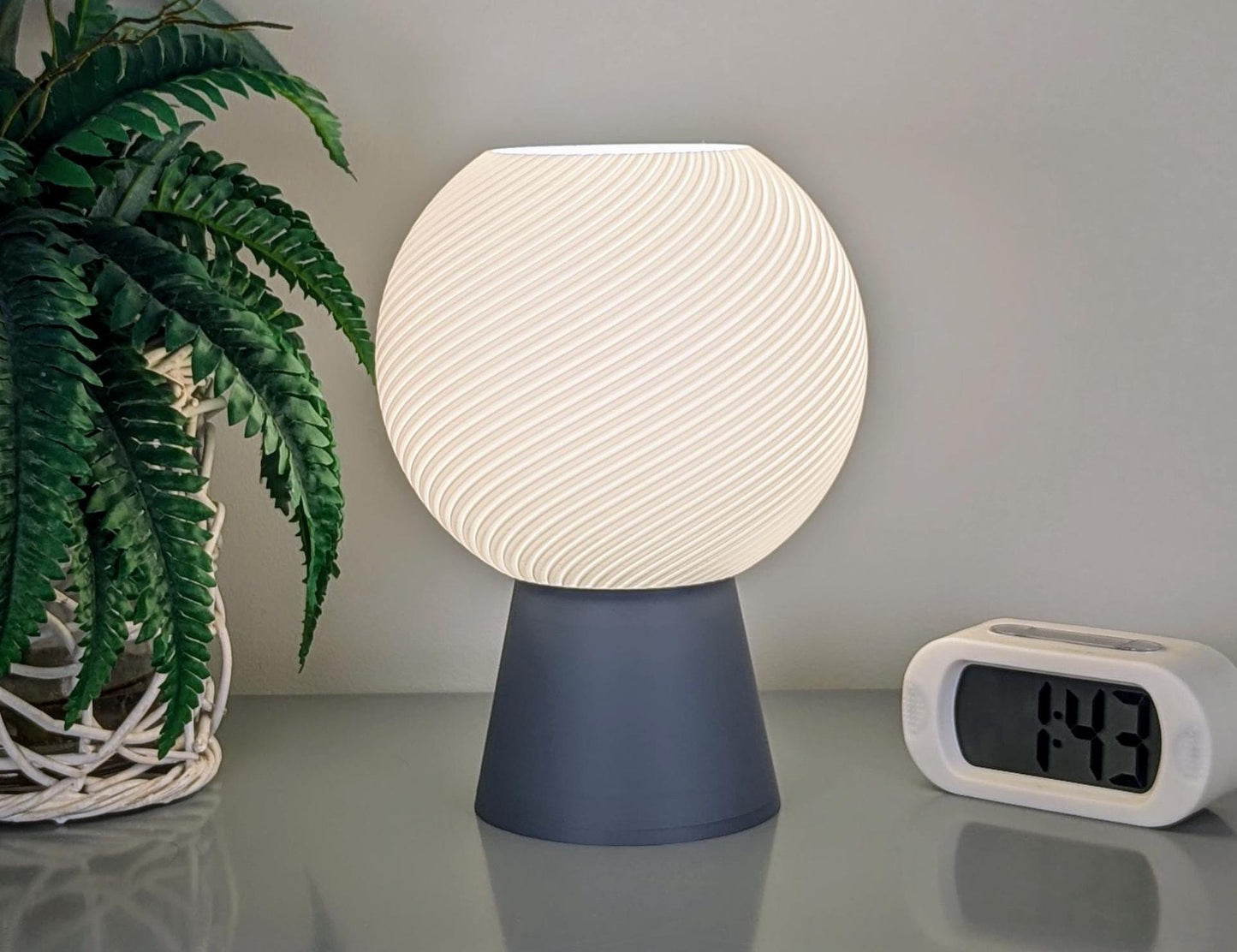 The Vortice Table Lamp by Dryft Home is a modern piece of lighting crafted with 3D printing technology. It features a swirling, textured spherical shade that emits a soft glow. Positioned on a reflective surface alongside a digital clock displaying 1:43 and accompanied by a fern in a woven basket, the lamp is made from biodegradable PLA plastic, emphasizing sustainable production.