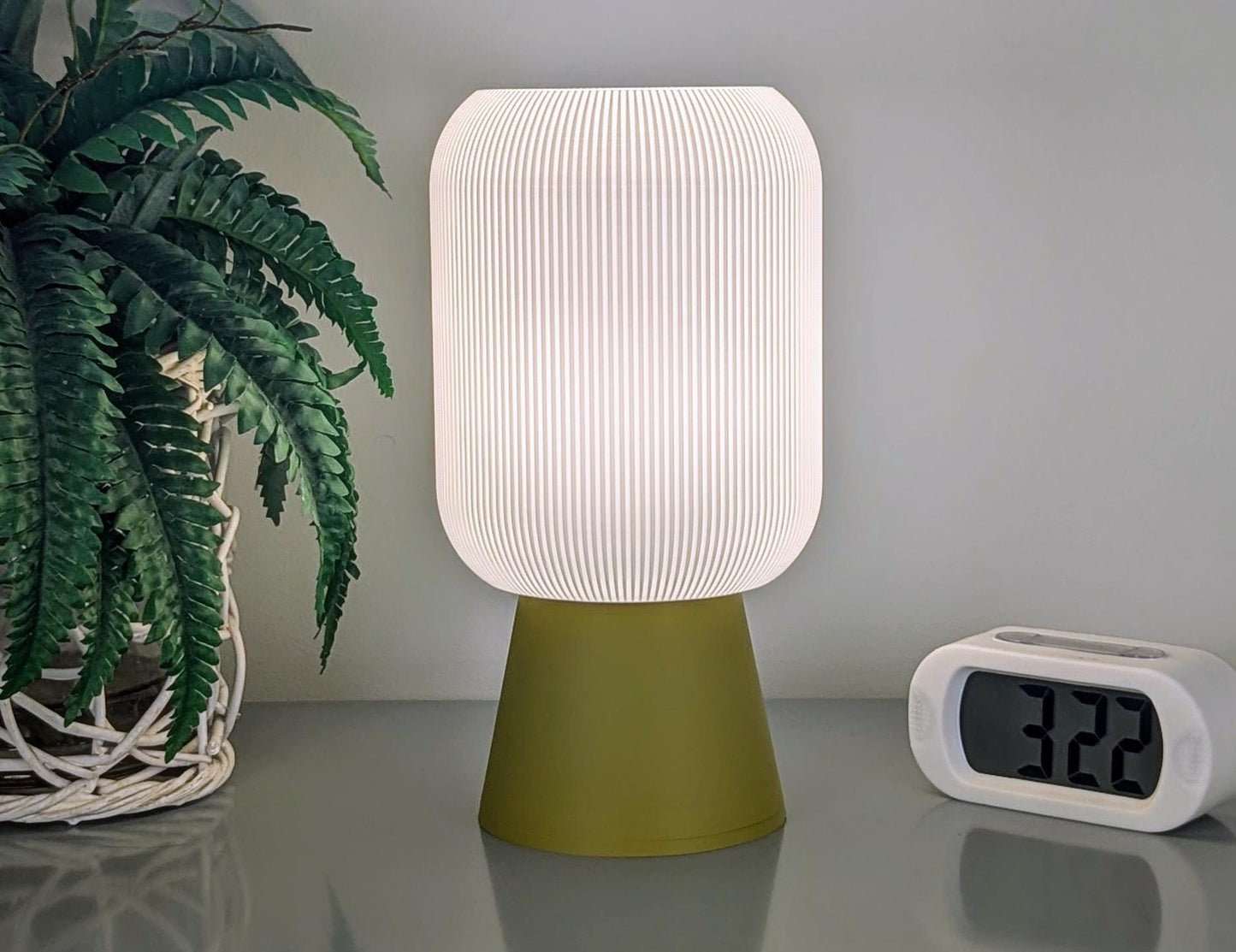 The Cumulis Table Lamp by Dryft Home, featuring a 3D printed pleated cylinder shade and an olive green base, made from eco-friendly materials, graces a reflective surface. Next to it, a digital clock shows 3:22 beside a potted plant with vibrant green leaves, showcasing eco-conscious design.