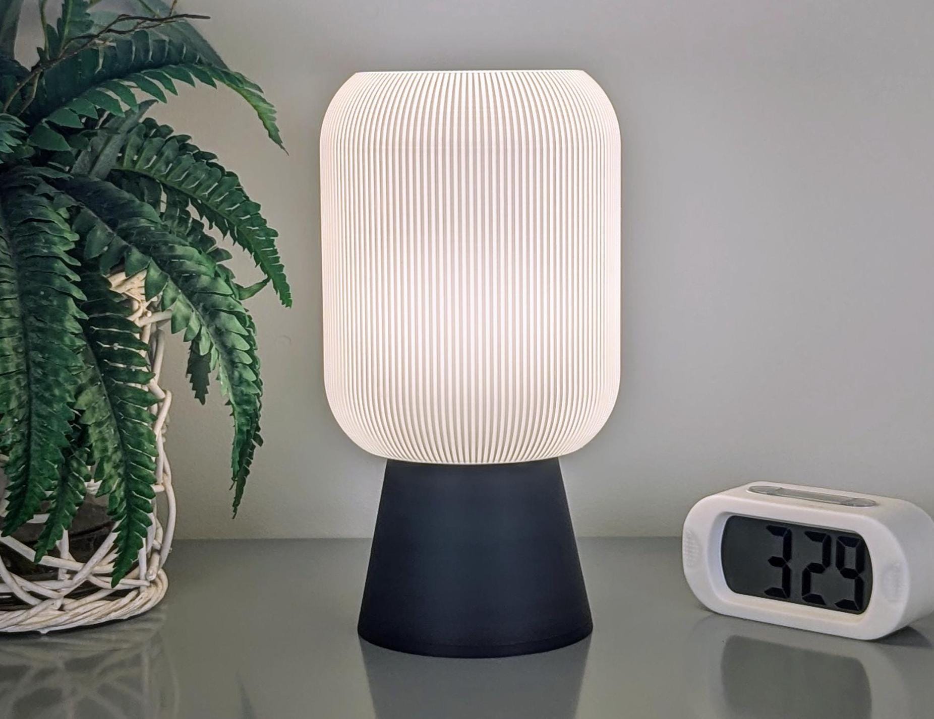 The Cumulis Table Lamp by Dryft Home, featuring a 3D printed pleated cylindrical shade in white and a black base made from sustainable materials, sits on a surface next to a digital clock displaying 3:29 and a potted plant with lush green leaves in a white basket.