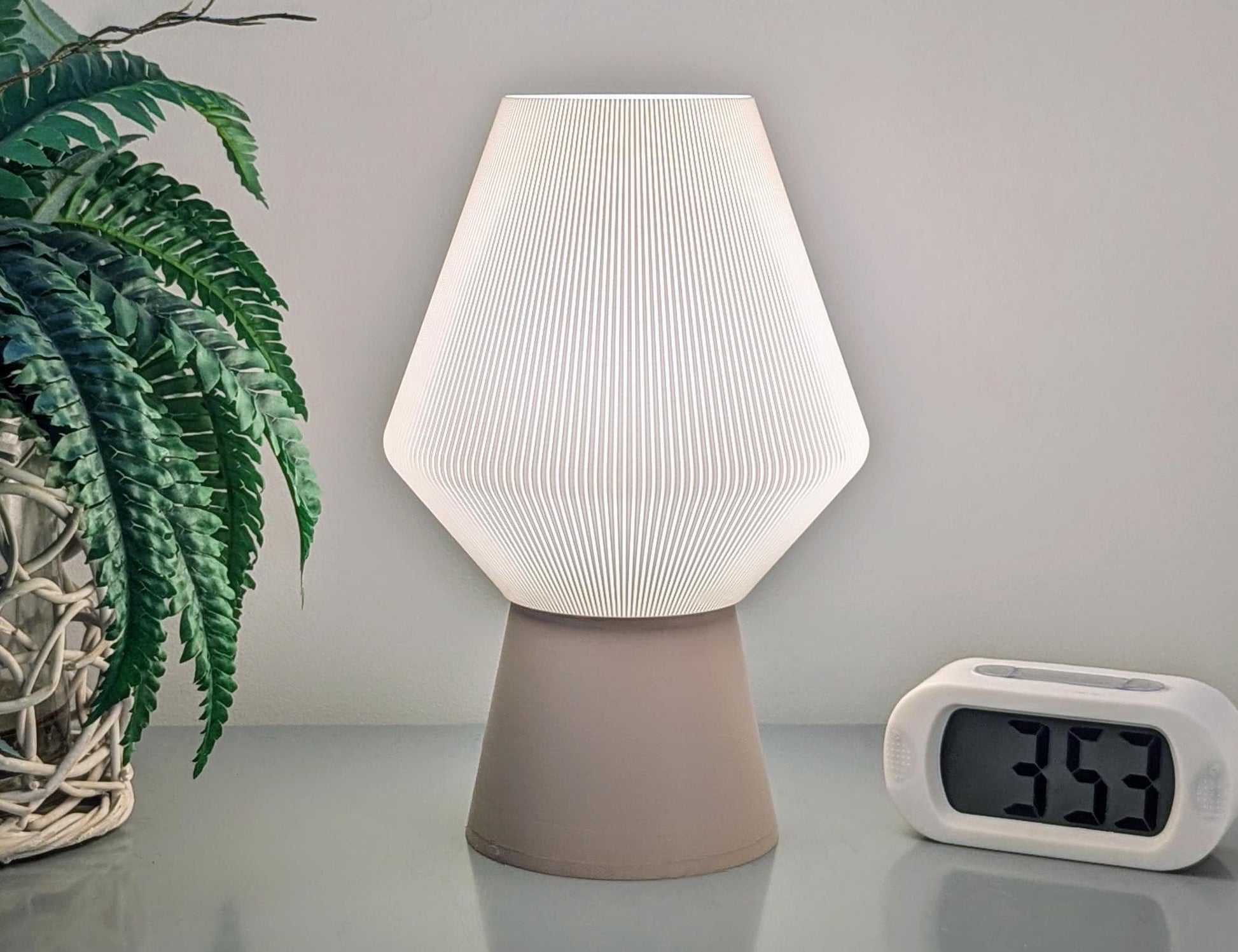 The Inversis Table Lamp by Dryft Home, featuring a modern geometric design and crafted from sustainable materials, offers a gentle glow. It sits alongside a white digital clock showing 3:53 and a potted plant with green leaves in a woven basket on a light-colored surface.