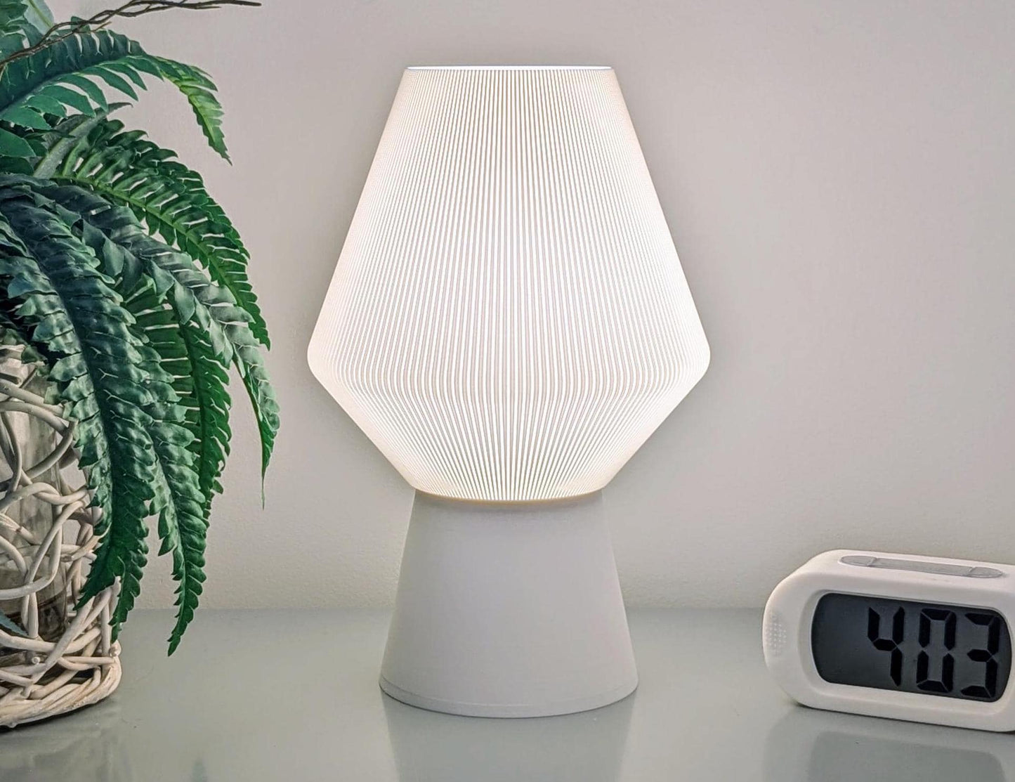 An Inversis Table Lamp by Dryft Home, featuring 3D-printed sculptural design and exuding a modern aesthetic, brightens up the space from its sleek resting place. On its left sits a woven basket housing a leafy green plant, while to the right stands a digital clock displaying 4:03 against an unadorned wall background.