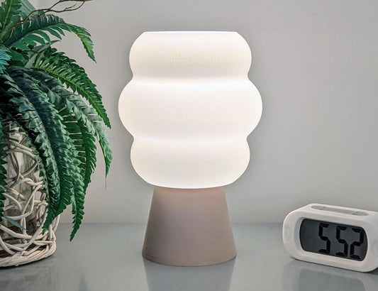 The Mammatus Table Lamp, a modern piece from Dryft Home featuring a wavy white shade and tan base crafted from environmentally friendly biodegradable PLA plastic, sits elegantly on a reflective surface. To its left is a potted plant with lush green leaves, while to its right stands a digital clock displaying 5:59. The scene is set against a plain gray wall backdrop.