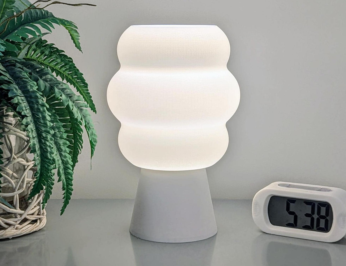 The Mammatus Table Lamp – 3D Printed Cloud-Inspired Lighting by Dryft Home, featuring a wavy design, sits on a table next to a digital clock displaying 5:38. Made from biodegradable PLA plastic, it enhances the scene with sustainable aesthetics alongside a potted fern adorned with wicker detailing on the left.