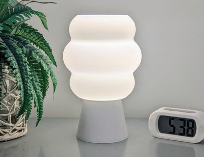 The Mammatus Table Lamp – 3D Printed Cloud-Inspired Lighting by Dryft Home, featuring a wavy design, sits on a table next to a digital clock displaying 5:38. Made from biodegradable PLA plastic, it enhances the scene with sustainable aesthetics alongside a potted fern adorned with wicker detailing on the left.