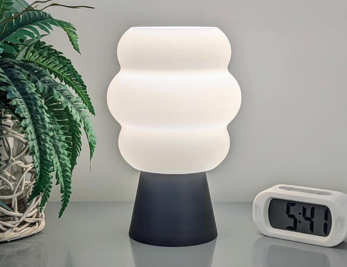 The Mammatus Table Lamp by Dryft Home, created with eco-friendly methods, showcases a textured white shade atop a dark base. It rests on a reflective surface next to a clock showing 5:41. To the left, a potted plant with green leaves is nestled in a woven basket.