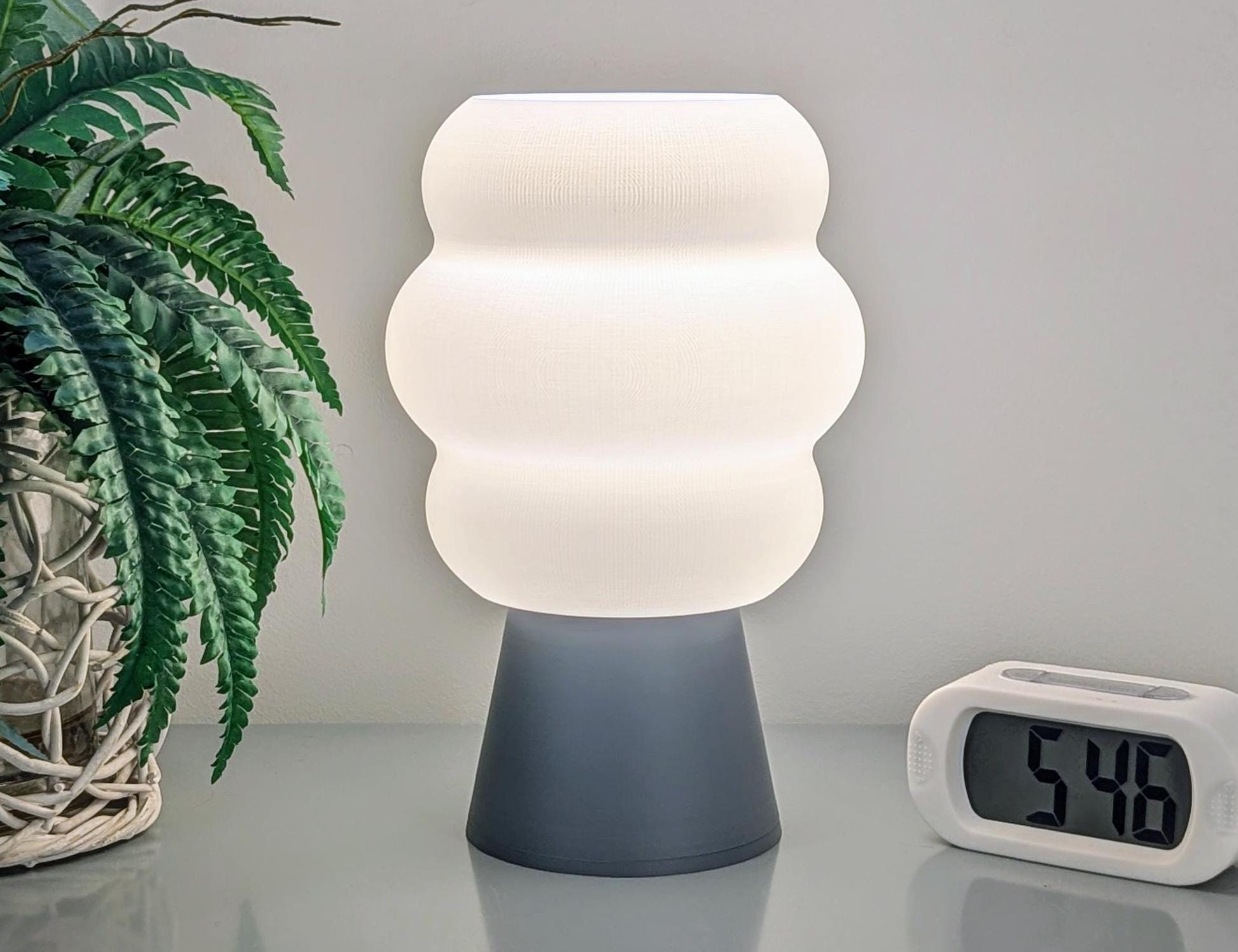 The Mammatus Table Lamp – a 3D Printed Cloud-Inspired Lighting piece by Dryft Home, is made from biodegradable PLA plastic and showcases a wavy white shade that casts a gentle glow across the table. Complemented by a digital clock displaying 5:48 and a potted plant in a woven basket, this arrangement highlights sustainable production while embodying a cozy, minimalist ambiance.