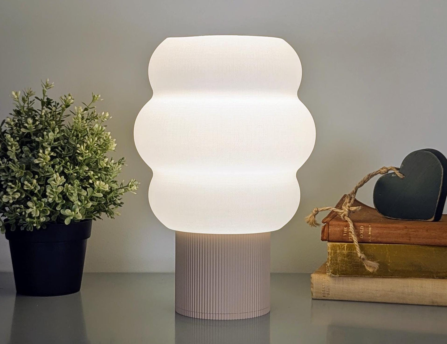 The Mammatus Table Lamp by Dryft Home, a 3D-printed cloud-inspired design in modern white biodegradable PLA, sits with a plant and twine-tied books topped with a heart ornament. Its wavy shade softly lit casts a warm glow, enhancing the cozy atmosphere.