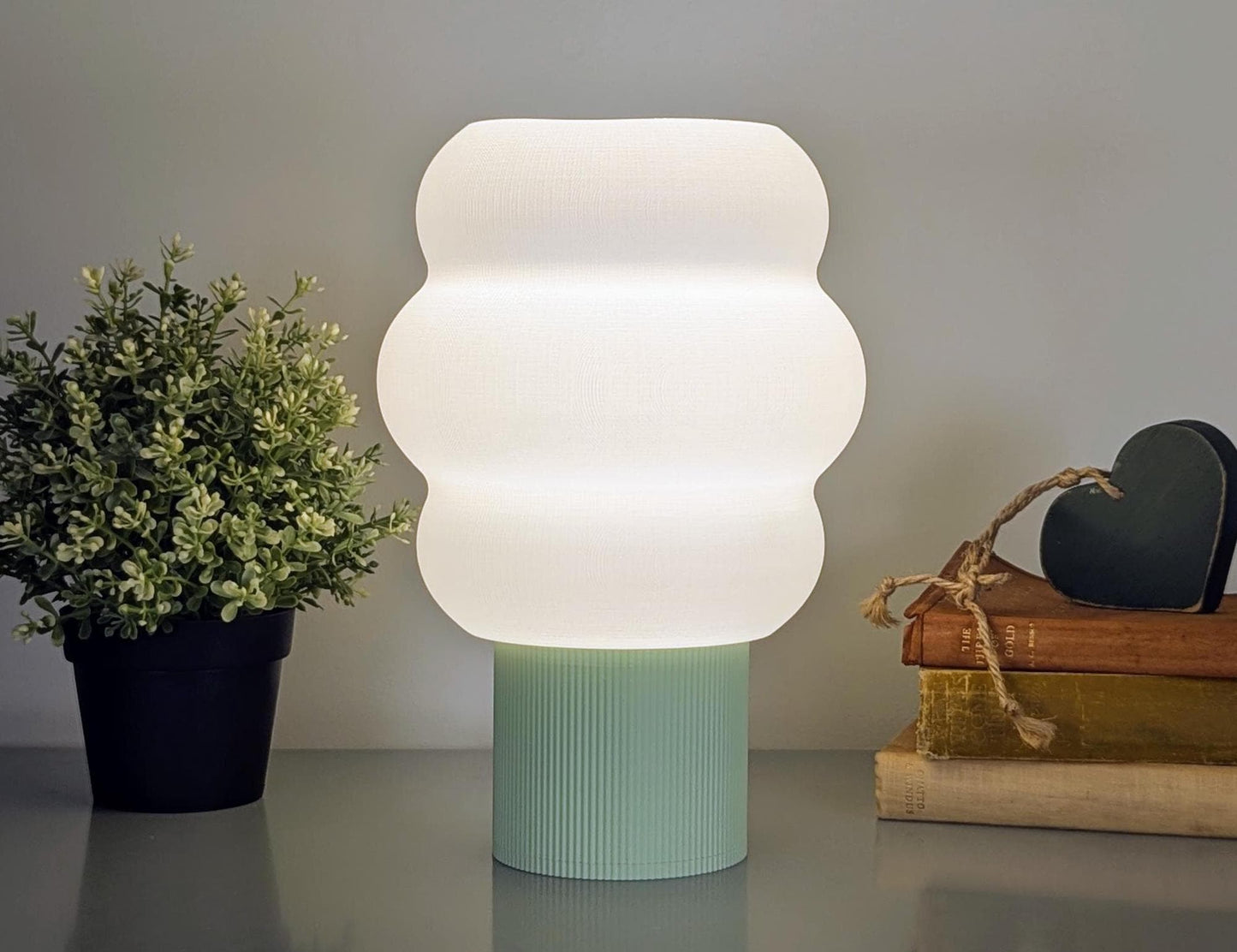 A Mammatus Table Lamp by Dryft Home, 3D printed and made from biodegradable PLA plastic, illuminates a table. To the left is a potted plant; to the right are stacked books and a small heart-shaped object. The plain gray wall highlights its sustainable production and cloud-inspired design.