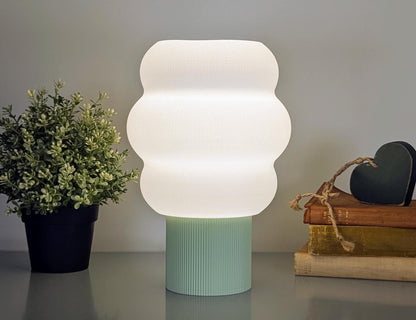 A Mammatus Table Lamp by Dryft Home, 3D printed and made from biodegradable PLA plastic, illuminates a table. To the left is a potted plant; to the right are stacked books and a small heart-shaped object. The plain gray wall highlights its sustainable production and cloud-inspired design.