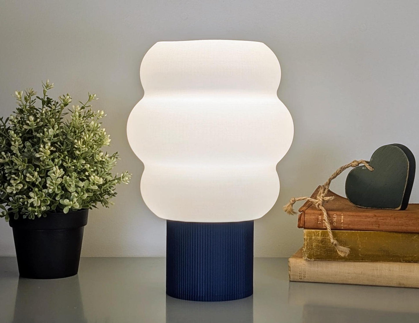 The Mammatus Table Lamp by Dryft Home, with its wavy white shade and dark blue base made from biodegradable PLA plastic, complements the nearby potted plant and stacked books, one adorned with a heart-shaped ornament. Its an eco-friendly, 3D printed cloud-inspired lighting solution.