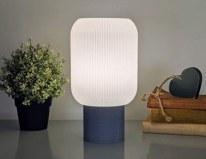 A Dryft Home Cumulis Table Lamp - 3D Printed Pleated Cylinder Lighting with a white textured shade, made from sustainable materials, is on the table. A potted plant sits to its left; on the right are stacked books and a decorative green heart with twine against a neutral gray wall.