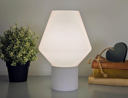 The Inversis Table Lamp by Dryft Home, with its 3D-printed geometric design, sits near a potted plant and books topped with a heart-shaped decoration. This modern white lamp emits a soft glow, adding sustainable decor flair and a cozy ambiance to the setting.