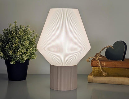 The Inversis Table Lamp by Dryft Home, featuring a 3D-printed geometric design, casts a soft glow over a table adorned with a small potted plant, books, and a heart-shaped ornament, creating a cozy ambiance that marries artistic technology with sustainable decor.