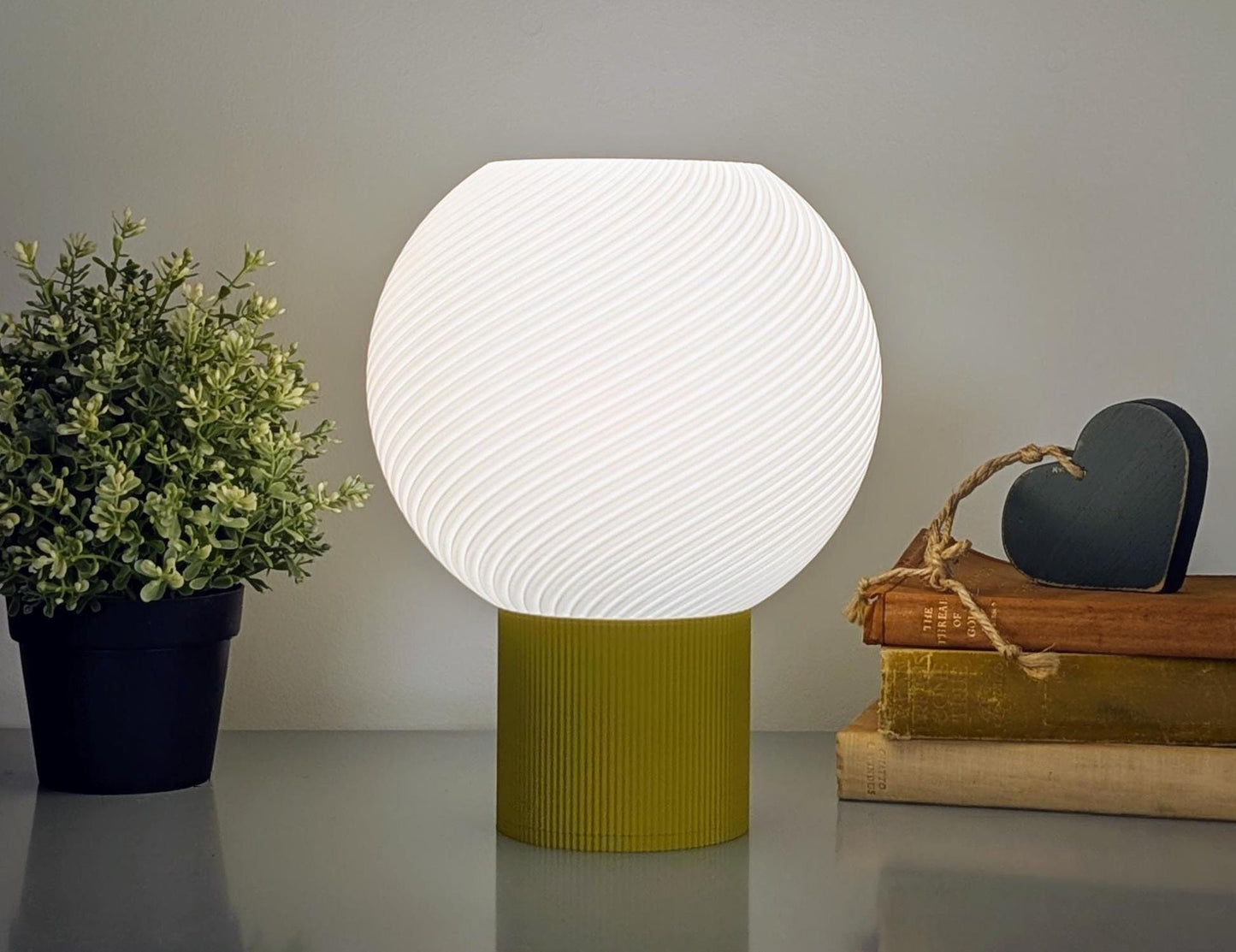 The Vortice Table Lamp from Dryft Home, a 3D printed spherical lamp with a textured surface, glows warmly on its yellow base. Accompanied by a small potted plant on the left and stacked books topped with a black heart-shaped ornament on the right, it creates a minimalist, cozy ambiance.