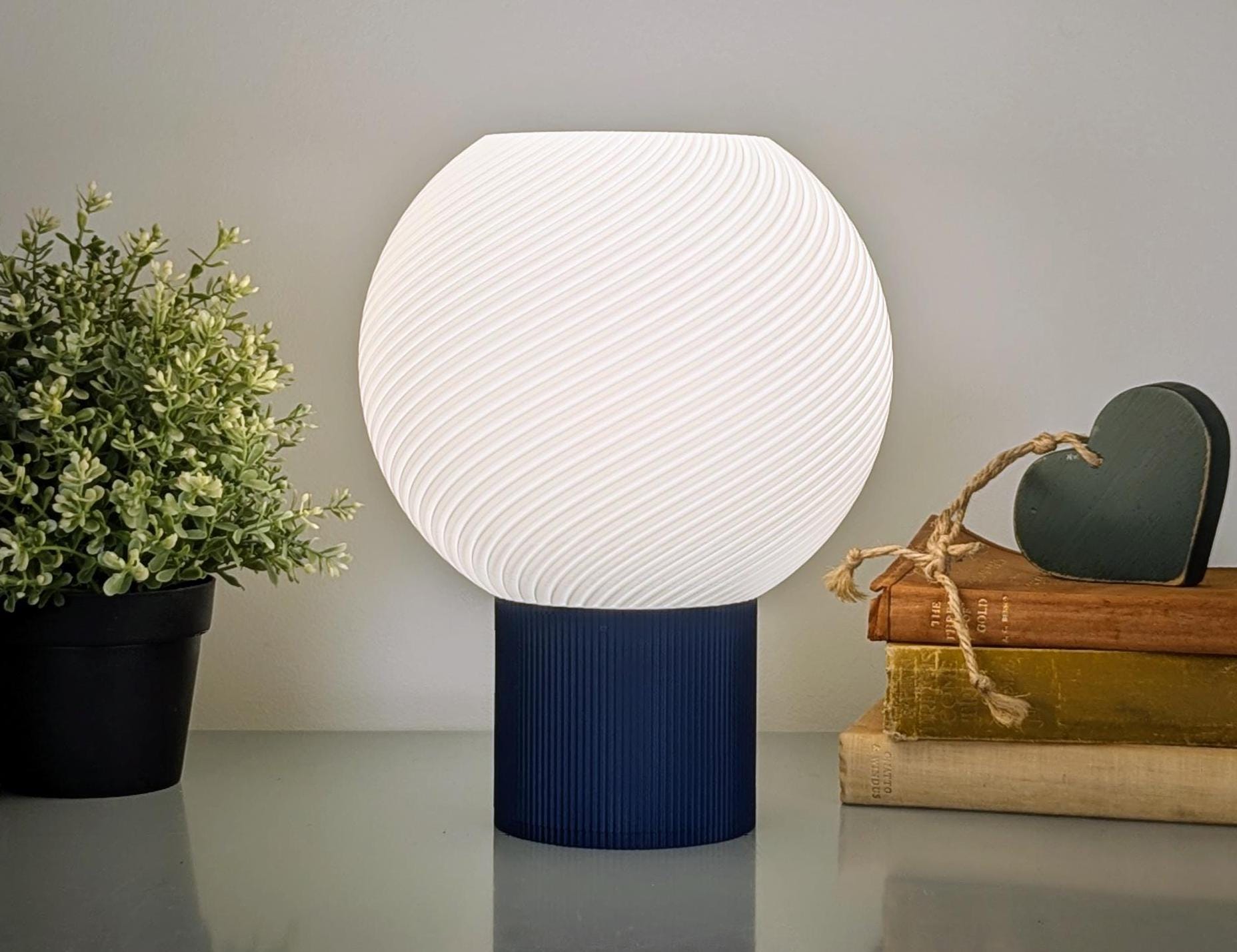 The Vortice Table Lamp by Dryft Home features a glowing white spherical shade made from sustainable materials and sits on a blue base. To its left is a potted plant, while to the right are stacked books and a heart-shaped ornament tied with twine.