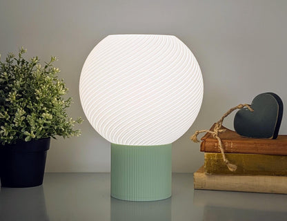 The Vortice Table Lamp from Dryft Home, crafted sustainably, showcases a glowing white sphere on a teal base. It complements a small potted plant and books tied with twine featuring a green heart ornament, all in minimalist style.