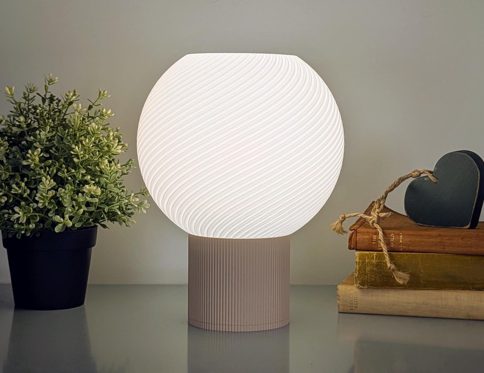 The Vortice Table Lamp by Dryft Home showcases a minimalist design with its textured, glowing spherical shade made from sustainable materials, elegantly placed beside a small potted plant and books topped with a decorative heart ornament.