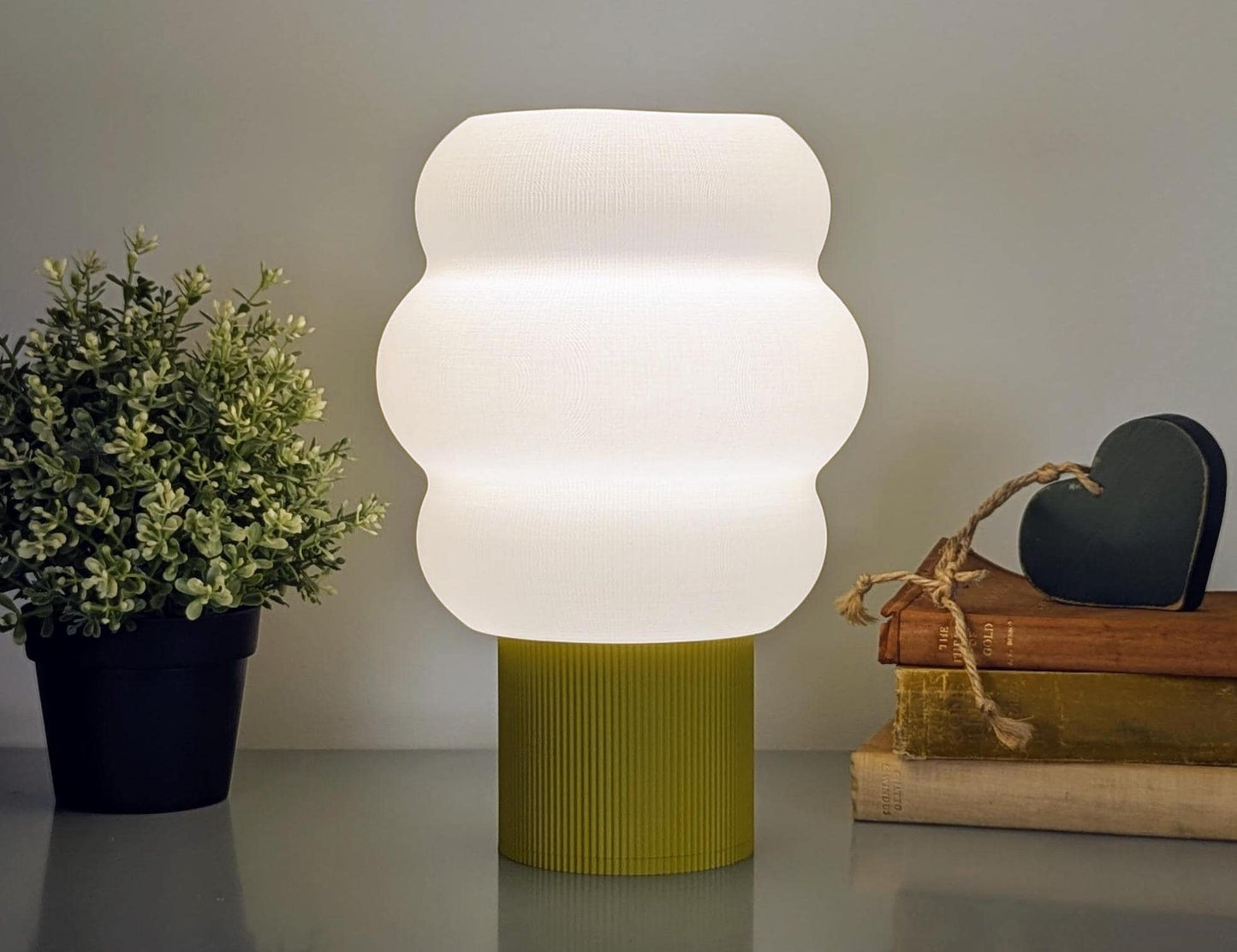 The Mammatus Table Lamp by Dryft Home features a modern, white 3D printed wavy design on a green base, crafted from biodegradable PLA plastic. Its set beside a small plant and stacked books, one tied with twine and topped with a wooden heart.