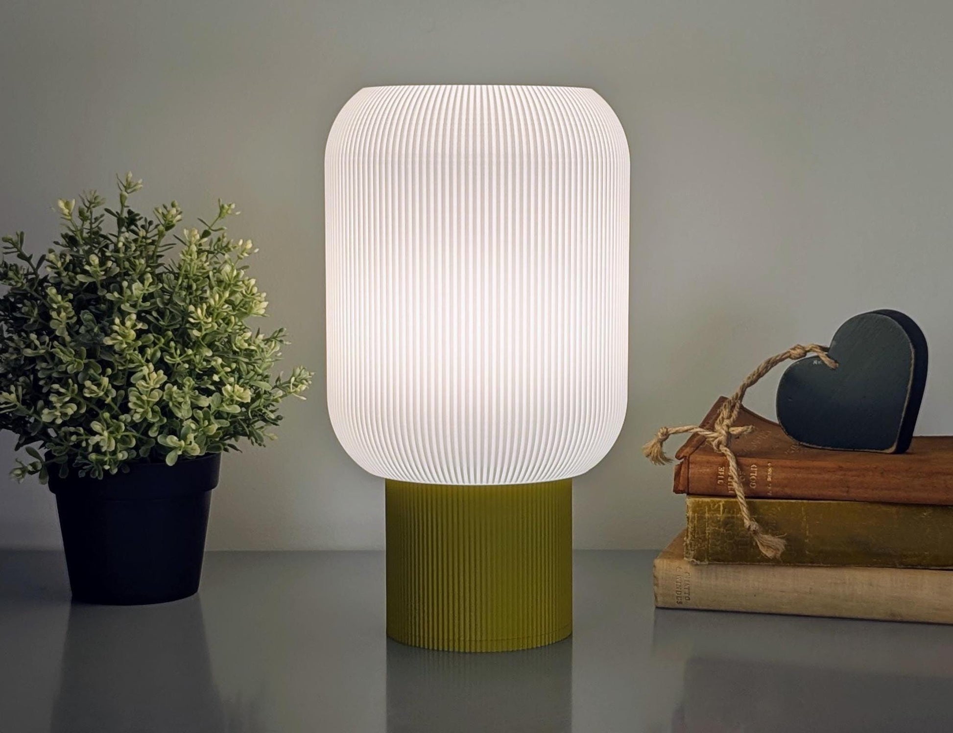 The Cumulis Table Lamp by Dryft Home, a 3D printed pleated cylinder lighting piece, is crafted from sustainable materials featuring a ribbed white shade and green base, casting an elegant glow beside a potted plant and books, topped with a dark heart-shaped ornament.